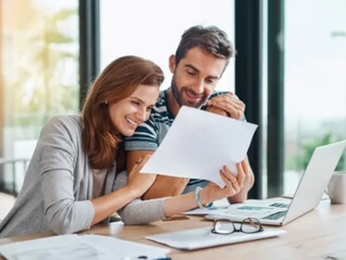 a man and a woman seated at a table with a laptop computer. The woman is holding a sheet of paper so the man can see it, and they are both smiling.