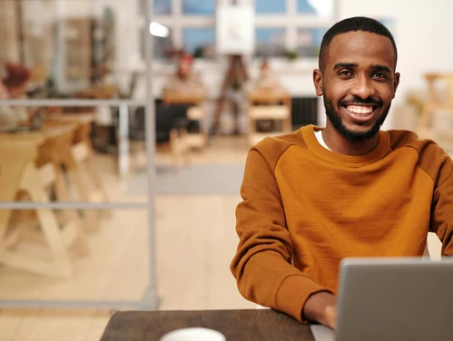 young man in rust colored shirt sitting at a desk looking at laptop