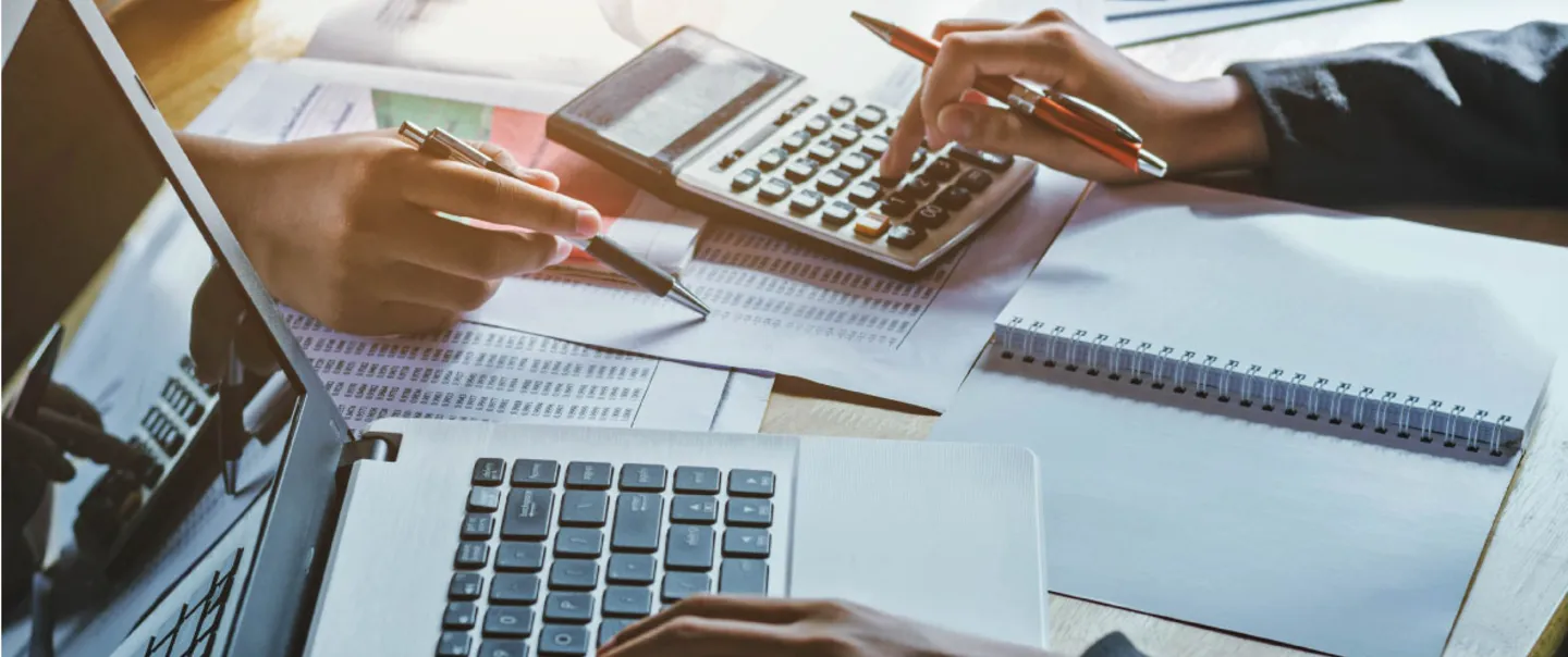 close-up photo of two people working across from each other at a desk. One person is using a calculator and a laptop computer, and the other is pointing to a piece of paper with a pen in their hand.