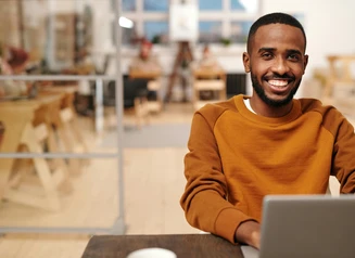 young man in rust colored shirt sitting at a desk looking at laptop